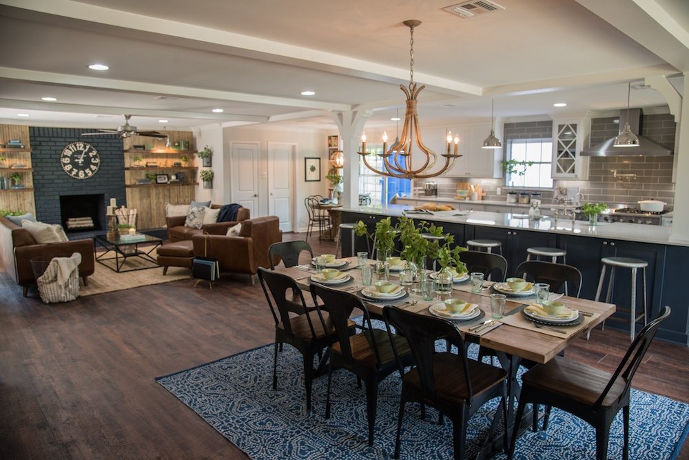 Dining table with black metal chairs in an open plan room, with a dark brick fireplace and the kitchen behind